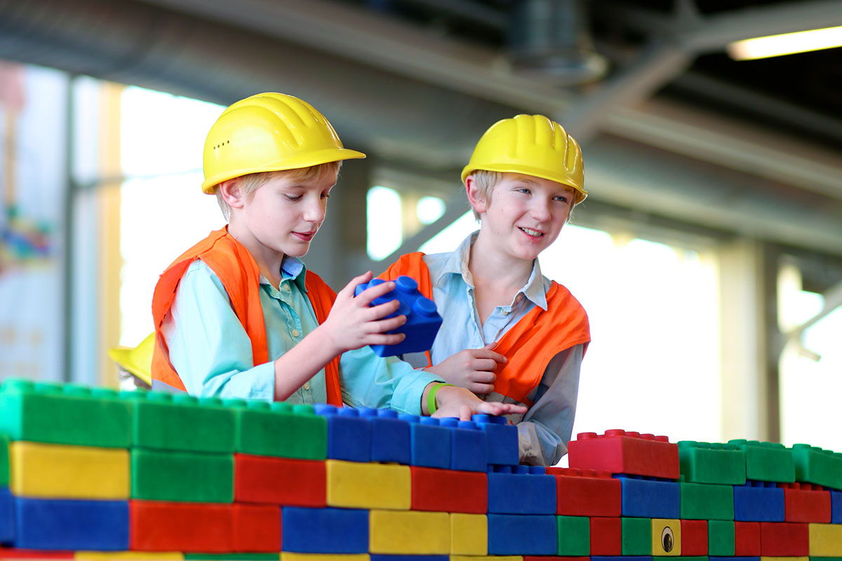 Two boys in safety helmets and high visibility jackets playing indoors. Schoolchildren building with construction bricks. Safety education for young kids. Playful work experience for future engineers.