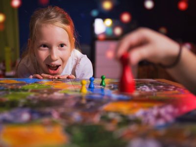 Board game concept - your move. Little girl watched the game and shock from the action move. Board game field, many figures. Kid girl play in board game at home on dark blurred background