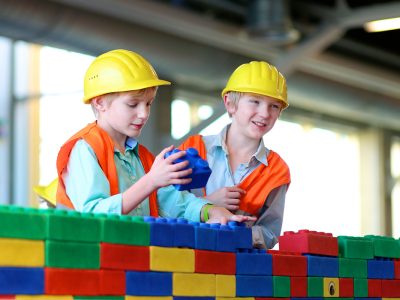 Two boys in safety helmets and high visibility jackets playing indoors. Schoolchildren building with construction bricks. Safety education for young kids. Playful work experience for future engineers.