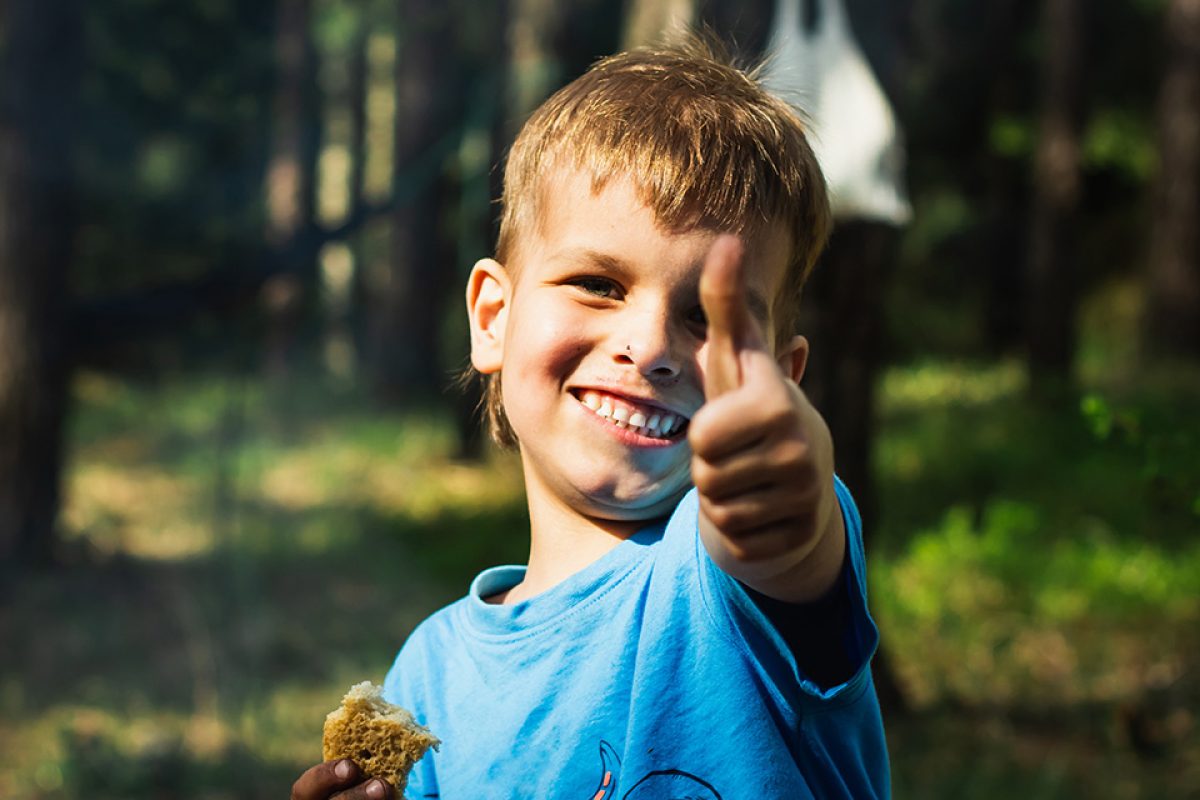 happy kid showing thumbs up sign on green forest background. smiling boy demonstrating optimistic gesture on sunny nature outdoors. happiness freedom childhood